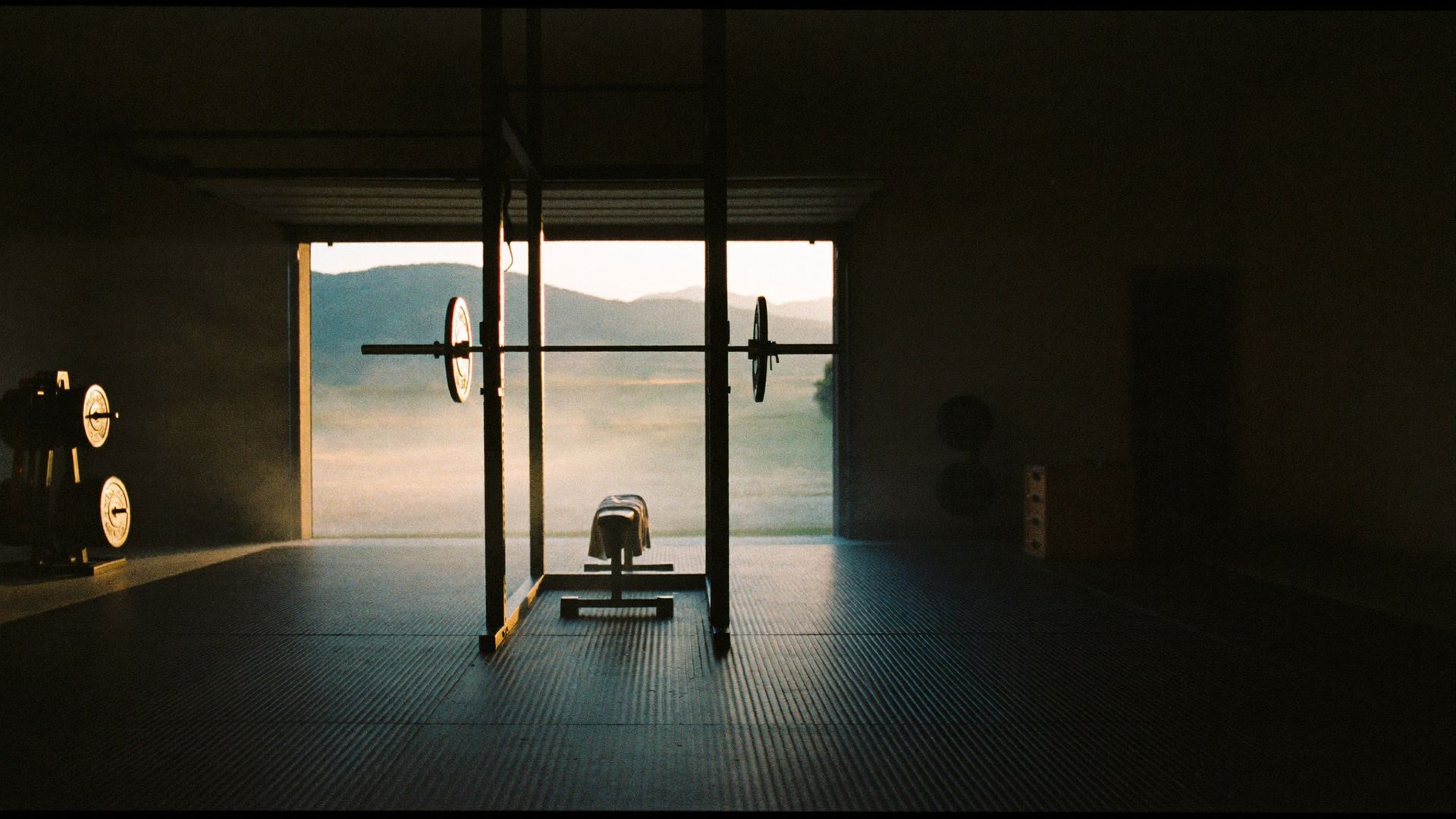 A garage gym at first light, the door part open, a barbell on a squat rack.