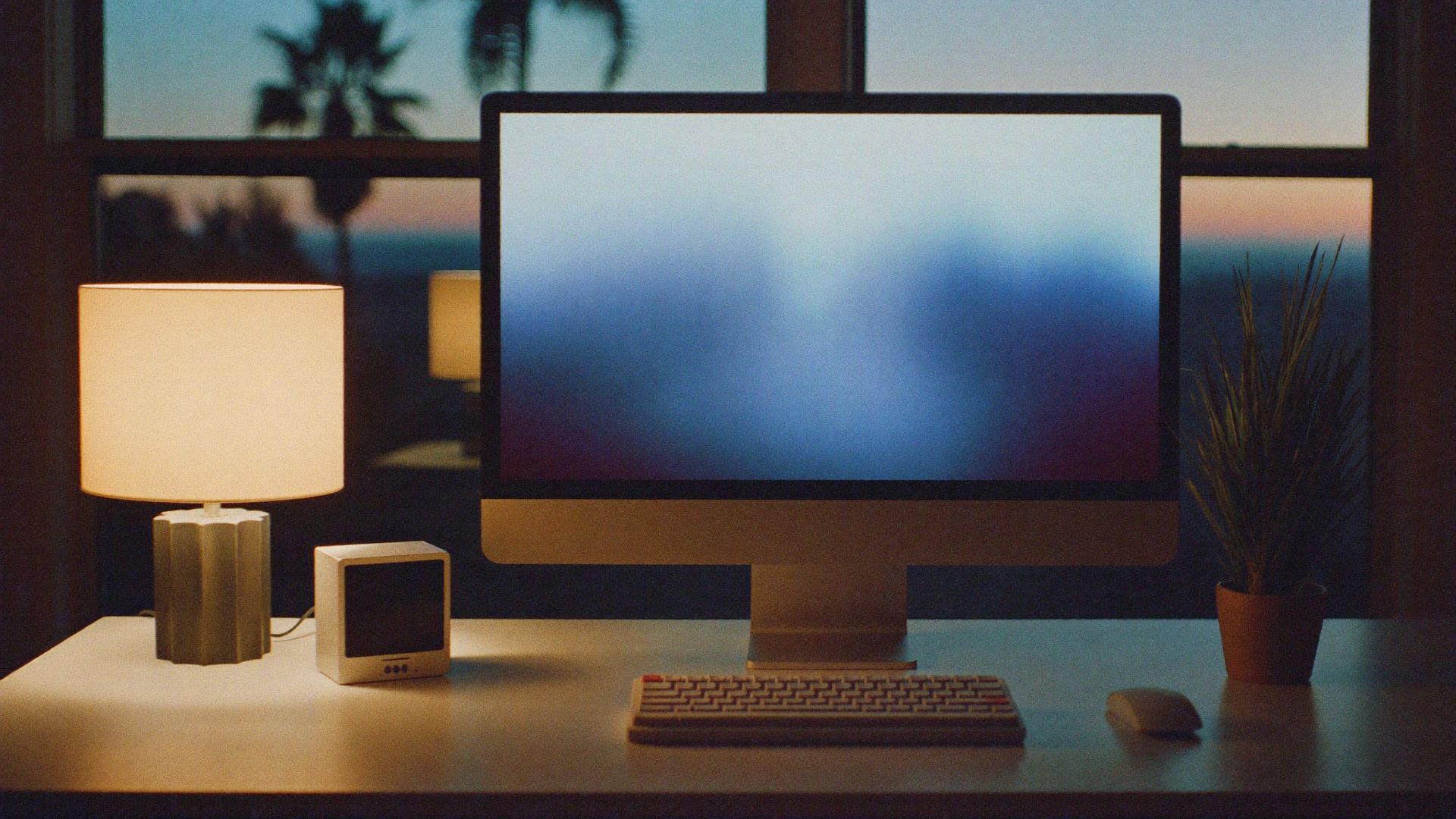 A home desk before sunrise, a monitor glowing blue, a small computer and a desk lamp.