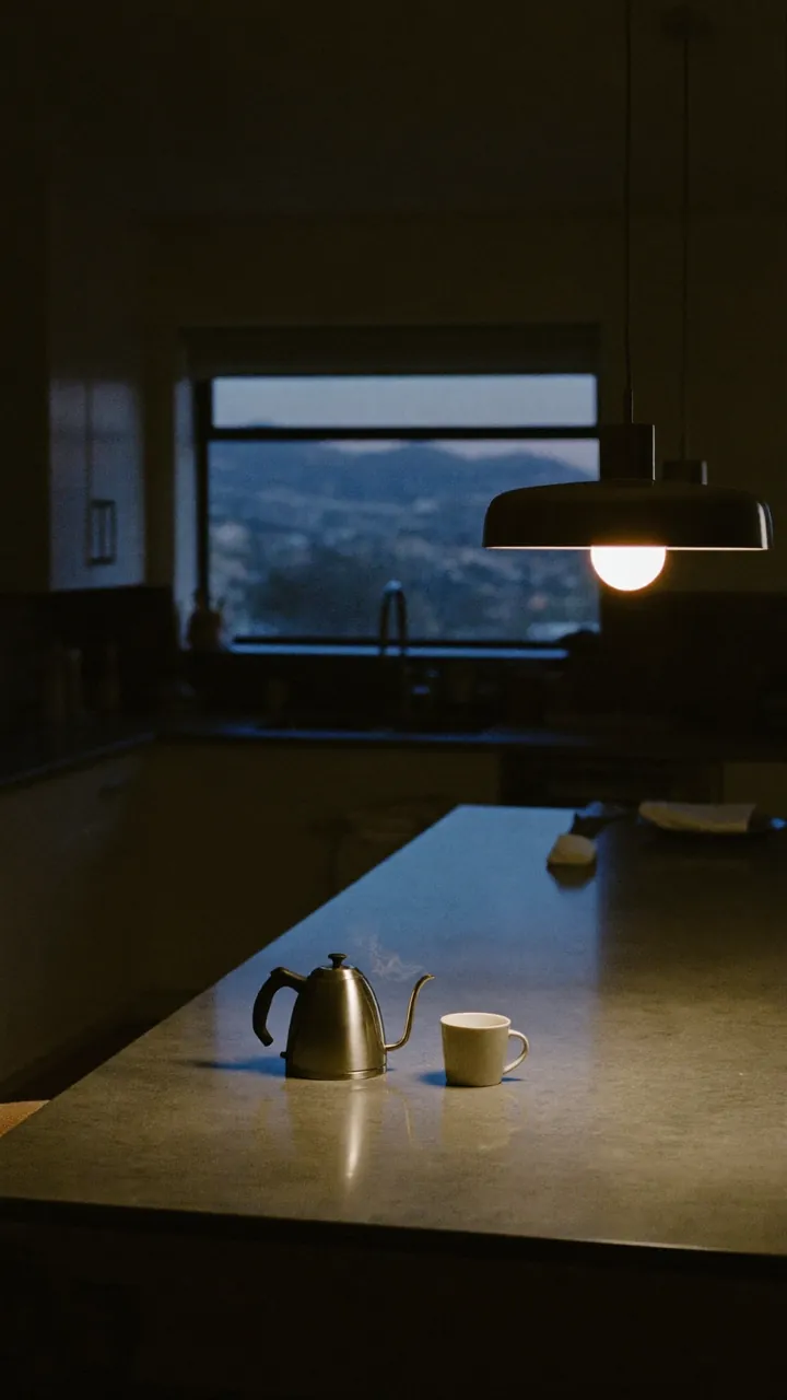A dark kitchen before sunrise, one pendant lamp over the island, coffee kettle and mug, hills in the window.